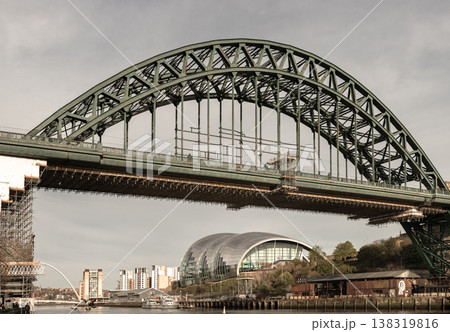 a striking industrial vista where the rugged strength of historic engineering meets the fluid curves of modern design. Dominating the upper frame is the iconic Tyne Bridge, its massive green steel 138319816