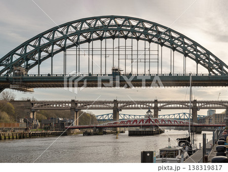 The Tyne Bridge green arch reigns over a horizon of engineering marvels. Its curve frames the historic High Level Bridge and the vibrant Swing Bridge reflected in the water. At the river edge, masts 138319817