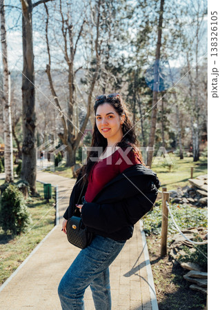 Young woman of slavic appearance posing on park pathway wearing red top jeans and black jacket holding handbag in sunny outdoor natural setting 138326105