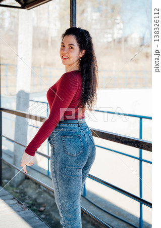 Full body portrait of smiling slavic woman with long curly dark hair wearing red top and blue jeans posing near railing in urban outdoor setting 138326112