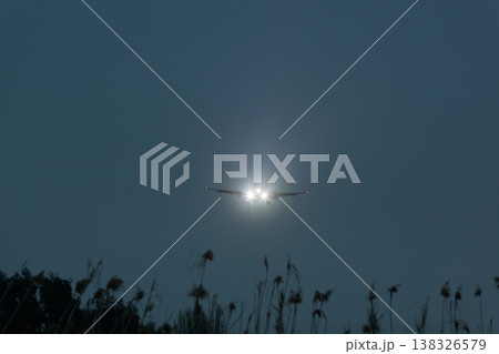 Airplane approaching landing with bright landing lights illuminated against a dark sky, silhouetted grass and vegetation in the foreground creating a dramatic scene Airplane approaching landing with bright landing lights illuminated against a dark sky, silhouetted grass and vegetation in the foreground creating a dramatic scene 138326579