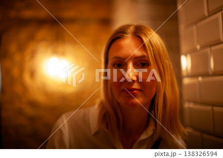 Female individual with blonde hair wearing a white shirt poses in a softly lit interior space with tiled walls and a warm light fixture in the background Female individual with blonde hair wearing a white shirt poses in a softly lit interior space with tiled walls and a warm light fixture in the background 138326594