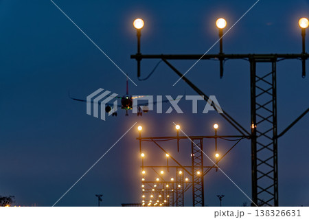 Airplane approaching landing with illuminated runway lights against a twilight sky, showcasing the airport's approach lighting system and surrounding infrastructure Airplane approaching landing with illuminated runway lights against a twilight sky, showcasing the airport's approach lighting system and surrounding infrastructure 138326631