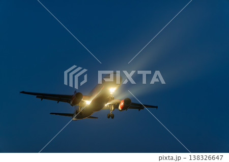 Airplane flying at night with landing lights illuminated, showcasing the aircraft's silhouette against a dark blue sky with faint clouds and atmospheric lighting effects 138326647