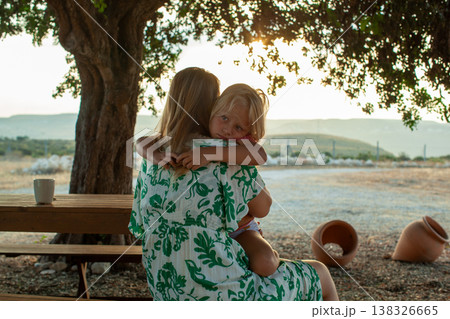 Woman in floral dress holds young child while sitting under a tree, with a scenic landscape and pottery visible in the background during golden hour Woman in floral dress holds young child while sitting under a tree, with a scenic landscape and pottery visible in the background during golden hour 138326665