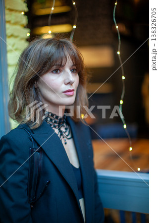 Female model with brown hair wearing a black blazer and choker stands near a window adorned with fairy lights in a cozy indoor setting Female model with brown hair wearing a black blazer and choker stands near a window adorned with fairy lights in a cozy indoor setting 138326705