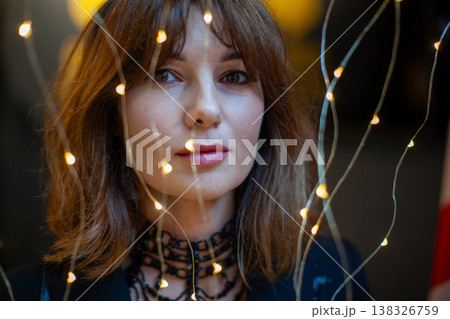 Female model with brown hair poses in front of decorative lights, soft focus background creates a warm ambiance, highlighting her features and attire 138326759