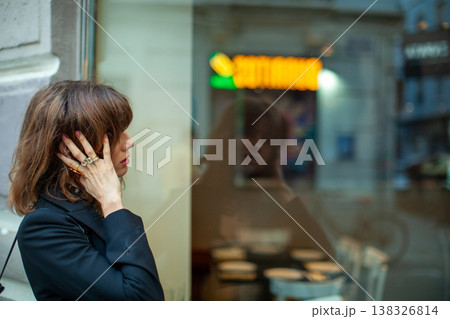 Female figure with brown hair wearing a black jacket stands outside a cafe, touching her ear while reflecting in the glass window with tables visible inside Female figure with brown hair wearing a black jacket stands outside a cafe, touching her ear while reflecting in the glass window with tables visible inside 138326814