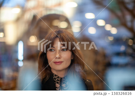 Female with brown hair and light makeup gazes thoughtfully through a glass window, with blurred lights and urban scenery visible in the background 138326816