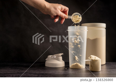 Protein drink. Hand pouring protein powder in a white shaker. Plastic tube on dark background. Body building, muscle growth and food supplement concept 138326860
