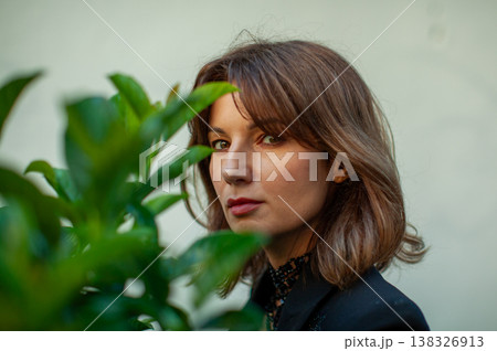 Female model with brown hair partially obscured by green foliage, gazing directly at the camera against a neutral background in an outdoor setting Female model with brown hair partially obscured by green foliage, gazing directly at the camera against a neutral background in an outdoor setting 138326913