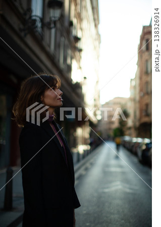 Young woman with brown hair wearing a black blazer stands on a city street, looking up towards buildings illuminated by sunlight in the background Young woman with brown hair wearing a black blazer stands on a city street, looking up towards buildings illuminated by sunlight in the background 138326914