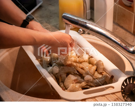 Woman washing fresh shrimp in kitchen sink. Preparation of raw seafood for meal cooking. Home culinary activity and healthy nutrition concept. Process of cleaning raw shellfish food ingredient. 138327340