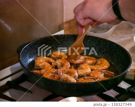 Man frying shrimp in a pan on gas stove. Process of cooking seafood with wooden spatula at home. Culinary preparation of healthy dish. Home lifestyle, kitchen leisure, nutrition concept. Man frying shrimp in a pan on gas stove. Process of cooking seafood with wooden spatula at home. Culinary preparation of healthy dish. Home lifestyle, kitchen leisure, nutrition concept. 138327349
