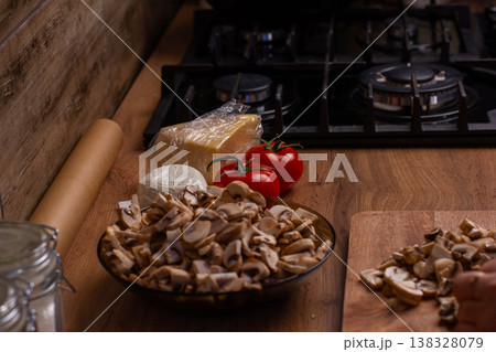 Fresh chopped mushrooms, whole tomatoes, and cheese blocks are ready on a wooden kitchen counter next to a gas stove, featuring a hand using a knife and a cutting board. Fresh chopped mushrooms, whole tomatoes, and cheese blocks are ready on a wooden kitchen counter next to a gas stove, featuring a hand using a knife and a cutting board. 138328079
