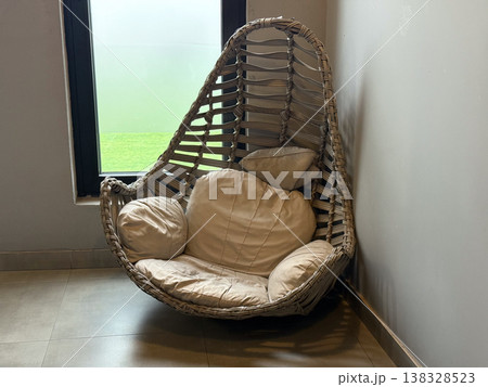 Wicker lounge chair with soft cushions placed near window in modern interior corner. Relaxation zone, waiting area seating, comfort furniture and interior styling concept 138328523