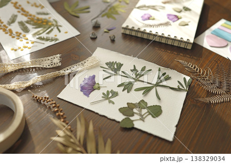 Creating botanical herbarium with pressed purple flowers and dried leaves in sketchbook. Creative workspace on wooden table with scissors, tweezers and crafting tools for DIY nature art project. Creating botanical herbarium with pressed purple flowers and dried leaves in sketchbook. Creative workspace on wooden table with scissors, tweezers and crafting tools for DIY nature art project. 138329034