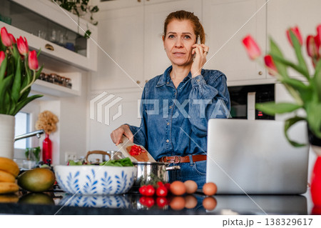 Mature woman making phone call while cooking in kitchen. Mature woman making phone call while cooking in kitchen. 138329617