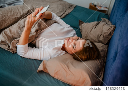 Middle aged woman using smartphone while lying in bed at home. Middle aged woman using smartphone while lying in bed at home. 138329628