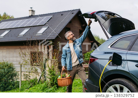 Man with basket with groceries charging eletric car. House with solar panels on roof behind. 138329752