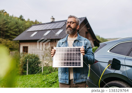 Man holding model of solar panel in hands. Charging electric car and house with solar panel system on roof behind him. Man holding model of solar panel in hands. Charging electric car and house with solar panel system on roof behind him. 138329764