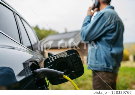 Man charging electric car in front of his house while making phone call. Man charging electric car in front of his house while making phone call. 138329766