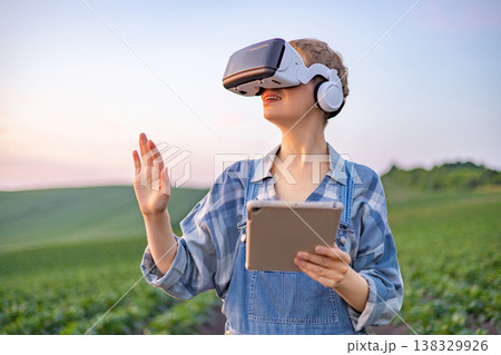 A person wearing a virtual reality headset and holding a tablet stands in a green field, exploring modern agricultural technology A person wearing a virtual reality headset and holding a tablet stands in a green field, exploring modern agricultural technology 138329926
