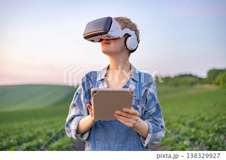 A person wearing a virtual reality headset and holding a tablet stands in a green field at sunset, symbolizing modern agricultural technology A person wearing a virtual reality headset and holding a tablet stands in a green field at sunset, symbolizing modern agricultural technology 138329927