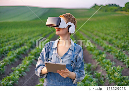 A young woman in overalls and a plaid shirt uses a tablet and VR headset while standing in a lush green field with rows of crops A young woman in overalls and a plaid shirt uses a tablet and VR headset while standing in a lush green field with rows of crops 138329928