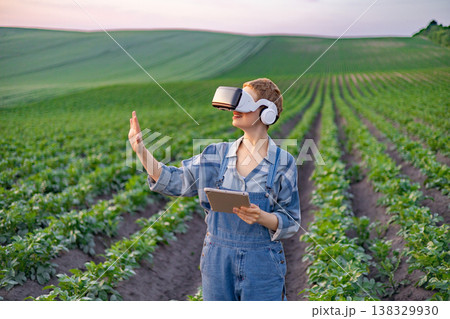 A person wearing a virtual reality headset and holding a tablet stands in a field of crops, exploring modern agricultural technology A person wearing a virtual reality headset and holding a tablet stands in a field of crops, exploring modern agricultural technology 138329930