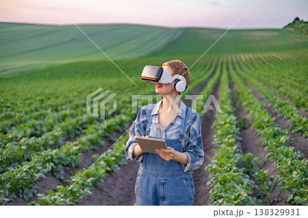 A woman in overalls and a plaid shirt uses a tablet and VR headset while standing in a field of crops, embracing agricultural technology A woman in overalls and a plaid shirt uses a tablet and VR headset while standing in a field of crops, embracing agricultural technology 138329931