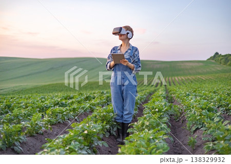A farmer wearing virtual reality goggles and headphones holds a tablet while standing in a field of crops, embracing agricultural technology A farmer wearing virtual reality goggles and headphones holds a tablet while standing in a field of crops, embracing agricultural technology 138329932
