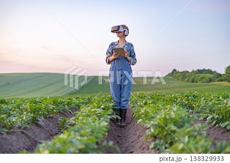 A person in overalls and VR headset stands in a field of crops holding a tablet, representing agricultural technology 138329933