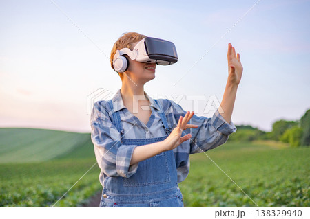 A young woman wearing overalls and a plaid shirt uses a virtual reality headset and headphones while standing in a lush green agricultural field at sunset 138329940