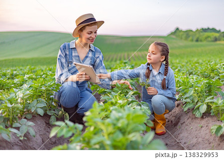 A woman and a young girl are examining plants in a field, with the woman holding a tablet 138329953