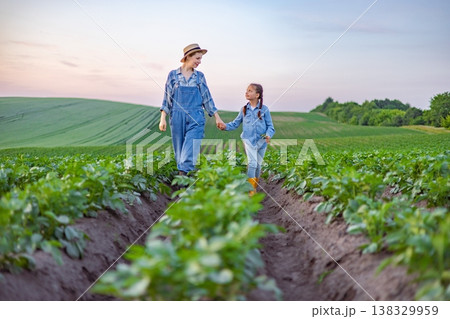 A mother and her young daughter walk hand-in-hand through rows of crops on a sunny day, enjoying the rural landscape and their connection to the land A mother and her young daughter walk hand-in-hand through rows of crops on a sunny day, enjoying the rural landscape and their connection to the land 138329959