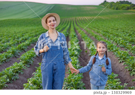 A woman and a young girl wearing denim overalls and straw hats stand hand-in-hand in a lush green field of crops 138329969