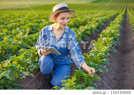 A woman in overalls and a hat kneels in a field, examining plants while holding a tablet computer 138330008