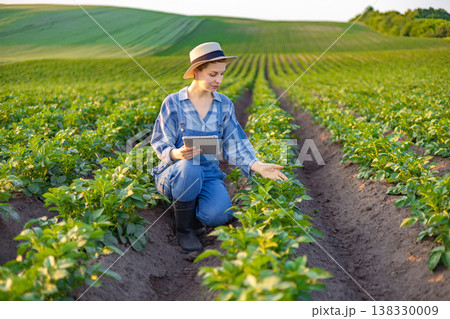 A woman in overalls and a hat examines crops in a field while holding a tablet computer 138330009