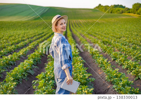 A woman wearing overalls and a hat stands in a field of crops holding a tablet computer A woman wearing overalls and a hat stands in a field of crops holding a tablet computer 138330013