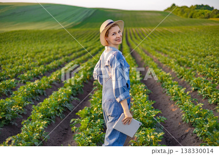A smiling woman wearing a straw hat and overalls stands in a lush green field holding a tablet, representing modern agriculture and technology A smiling woman wearing a straw hat and overalls stands in a lush green field holding a tablet, representing modern agriculture and technology 138330014