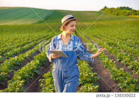 A woman in overalls and a hat uses a tablet computer while inspecting crops in a vast agricultural field during golden hour 138330022