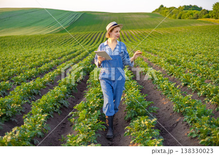 A woman in overalls and a hat walks through a potato field holding a tablet and gesturing with her hand A woman in overalls and a hat walks through a potato field holding a tablet and gesturing with her hand 138330023