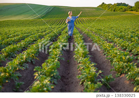 A woman in overalls and a hat walks through rows of potato plants, holding a tablet and gesturing towards the field 138330025