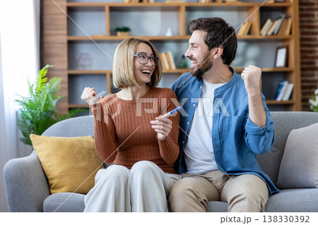 Cheerful young couple on a sofa celebrating a positive pregnancy test, embracing and smiling as they share joyful anticipation of starting a family and a new life together 138330392