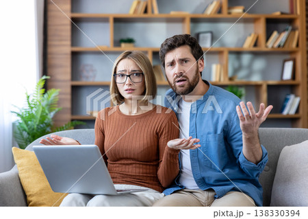 Young couple looking puzzled and shrugging while experiencing technical issues or an error on a laptop computer, sitting on a sofa in their living room Young couple looking puzzled and shrugging while experiencing technical issues or an error on a laptop computer, sitting on a sofa in their living room 138330394