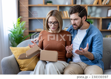 Couple sitting on a couch at home, looking frustrated and expressing disappointment after opening a damaged brown carton box from an online purchase or delivery 138330397