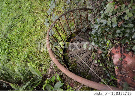A rusty spiral metal staircase surrounded by dense greenery and overgrown plants in Busaco Park, Portugal. Themes explore decay, abandonment, and the return of nature to man-made structures. 138331715