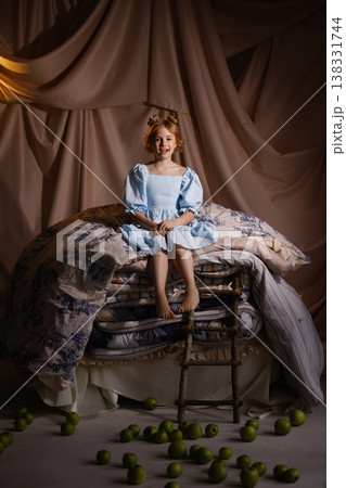 Little girl in blue dress sitting on stacked mattresses with apples, princess concept, studio drapery 138331744