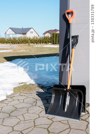 Two snow shovels leaning against a garage with melting snow, ready for winter cleanup. 138331789